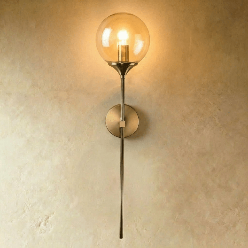 Brass wall sconce with clear glass sphere shade and long stem against gray wall, lifestyle setting with bed and plant.
